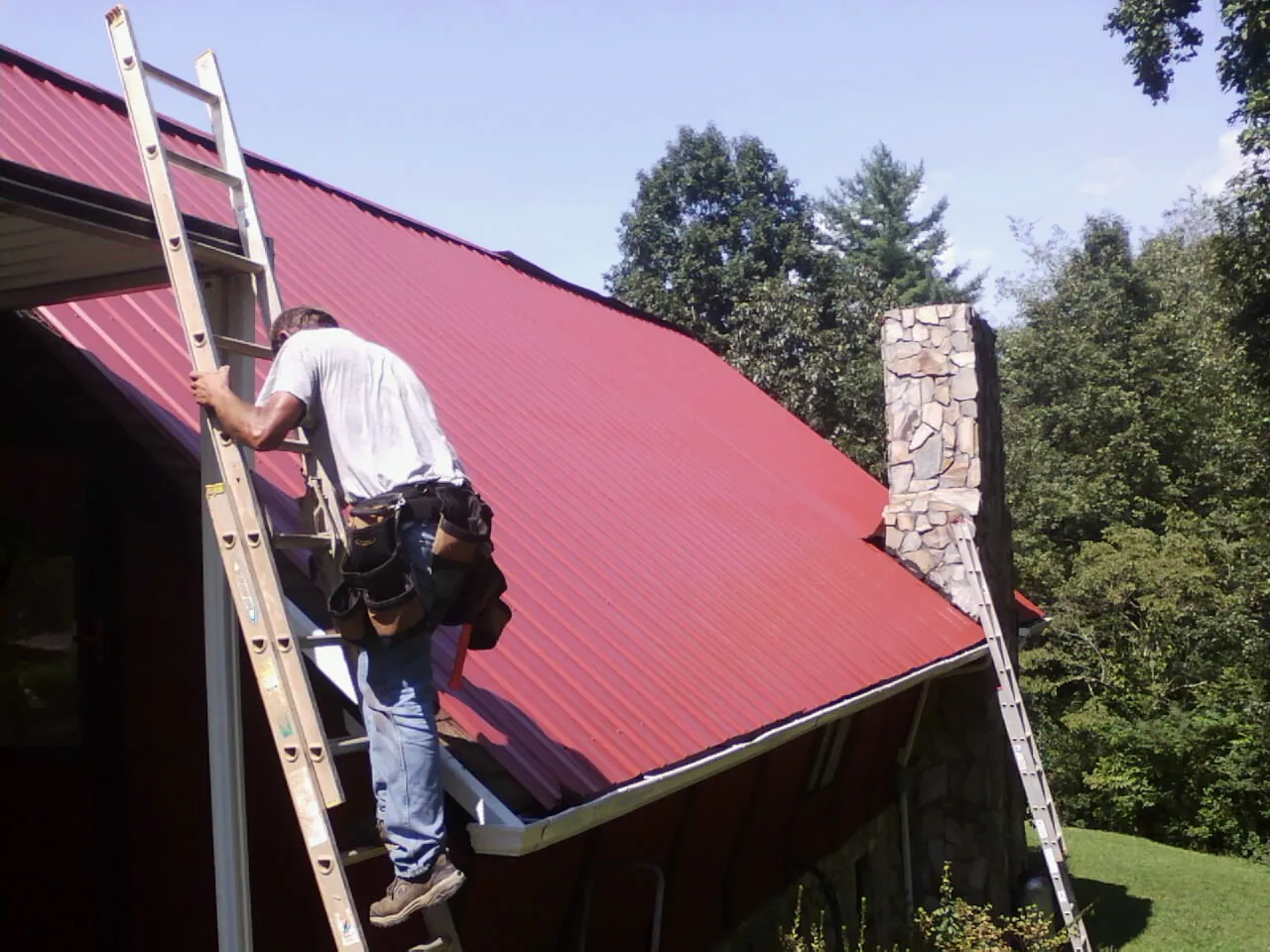Roofer on a ladder beside a red metal roof and stone chimney — Soffit & Fascia Repair by Roofing Brothers in Hydes
